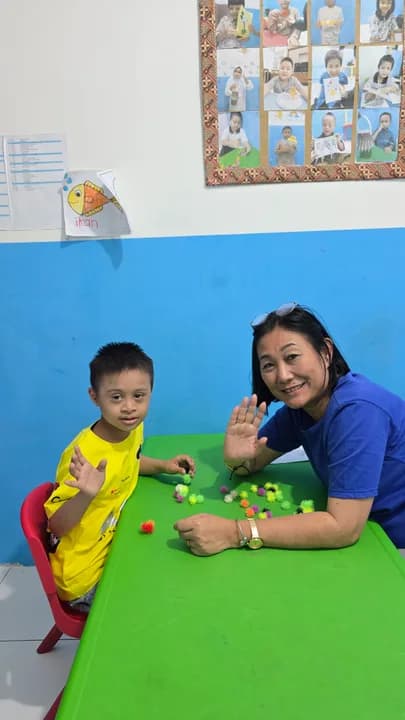 Psikolog tersenyum di samping anak berbaju kuning di meja hijau