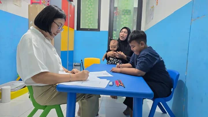 Psikolog mencatat di meja biru sementara seorang ayah mendampingi anaknya